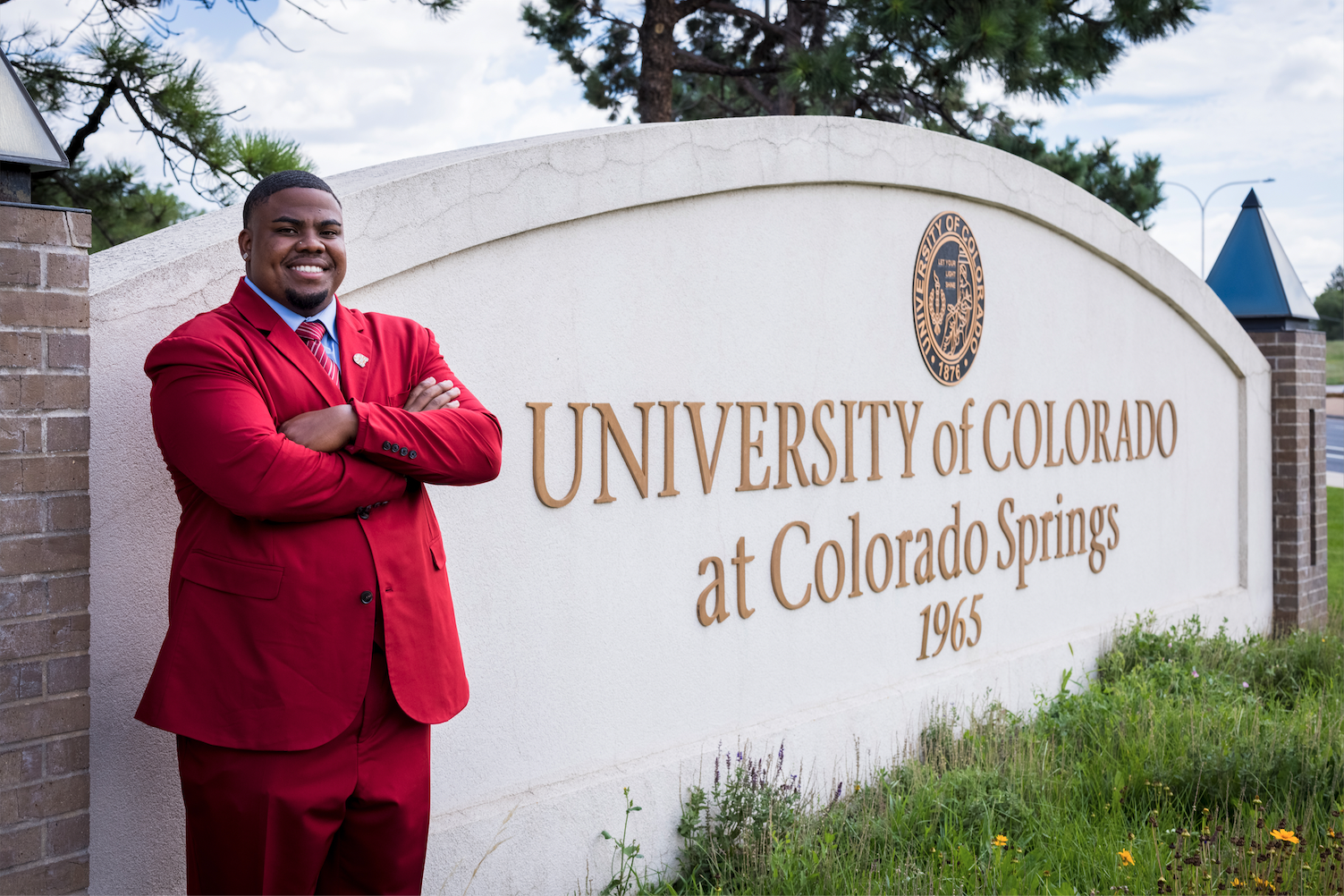 Michael "Axel" Brown wears a red suit and stands next to the University of Colorado Colorado Springs sign