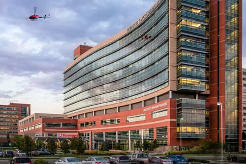 A twilight photo of the University of Colorado Hospital building with a helicopter in the sky and cars in the parking lot