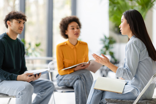 Two female and one male student sit on chairs with notebooks on their laps and have a conversation