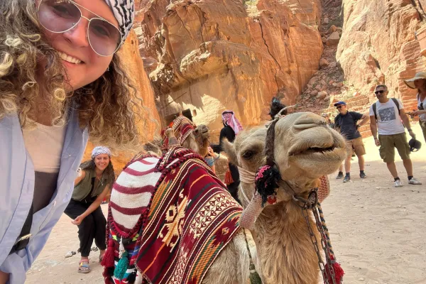 A photo of Sara Khammash wearing sunglasses and posing with a camel in a desert setting