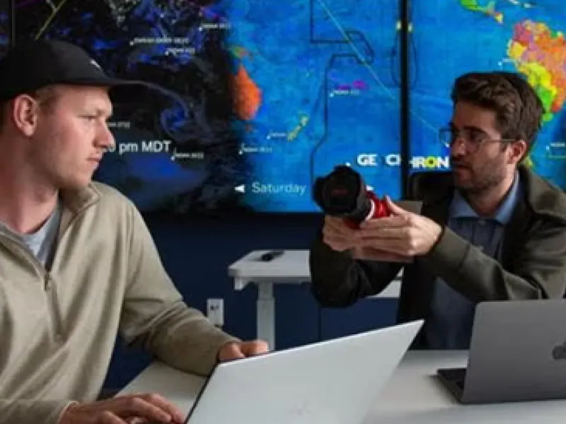 Two men are in front of a large world map, the one on the right is holding a camera