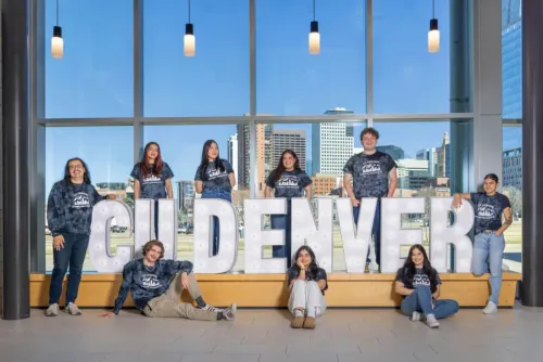 Students in blue shirts and blue jeans stand around a CU Denver sign